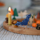 A Grimm's wooden bat decorative figure placed on a natural Grimm's celebration ring. A lit candle and other decorative items can be seen placed on the ring in the background. 
The decorative figures are part of a wide range of figures and rings available here at Babipur.