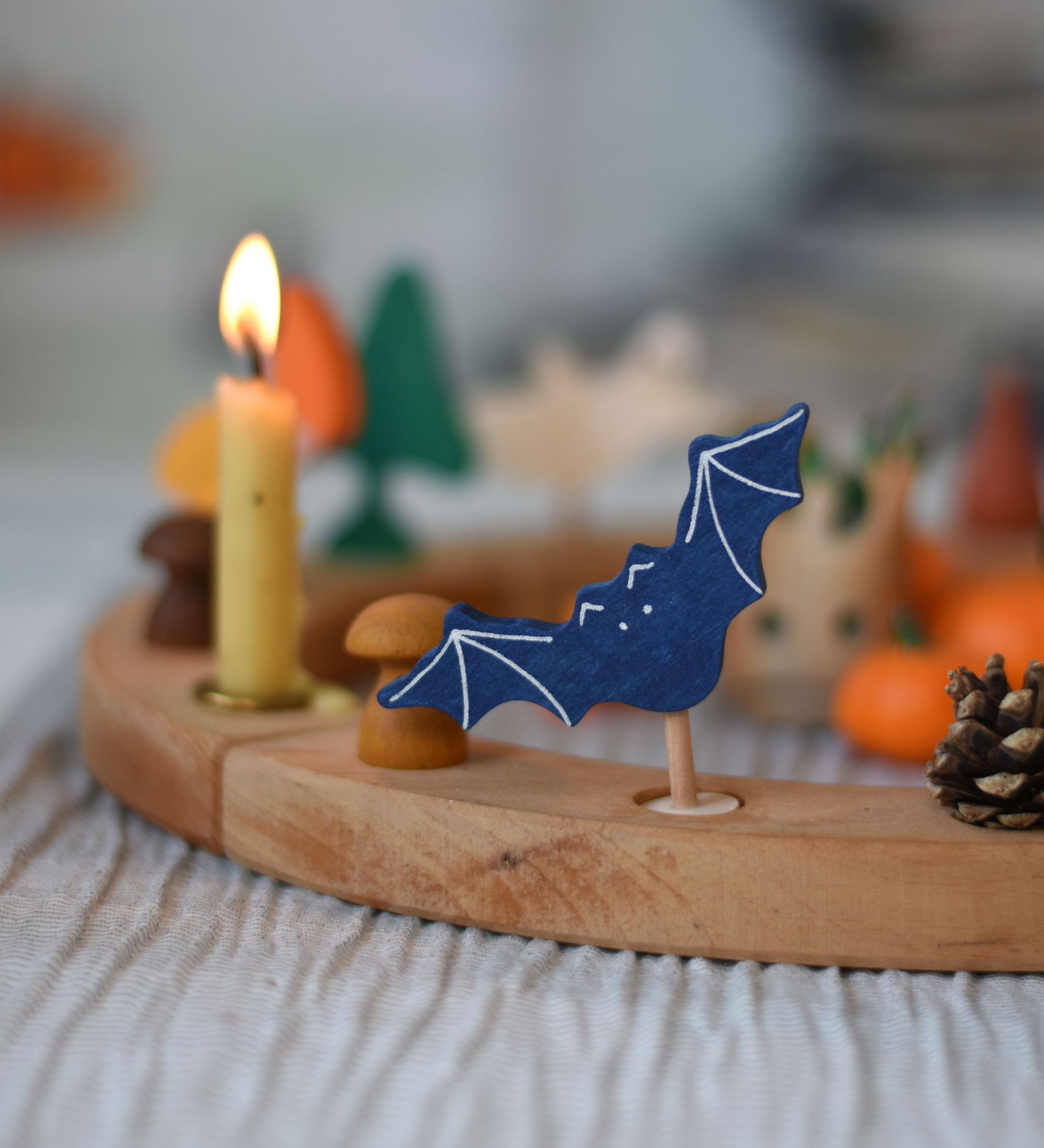 A Grimm's wooden bat decorative figure placed on a natural Grimm's celebration ring. A lit candle and other decorative items can be seen placed on the ring in the background. 
The decorative figures are part of a wide range of figures and rings available here at Babipur.