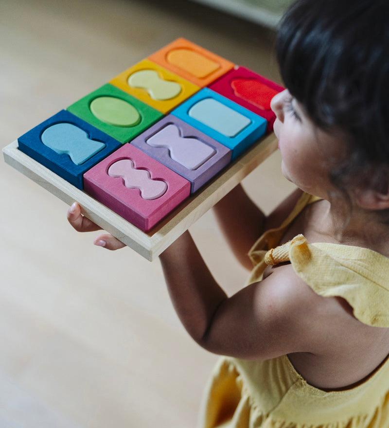 A child holding the Grimm's windows building set blocks set on the floor in front of them. These Grimm's toys are part of a wide range of wooden toys available here at Babipur. 