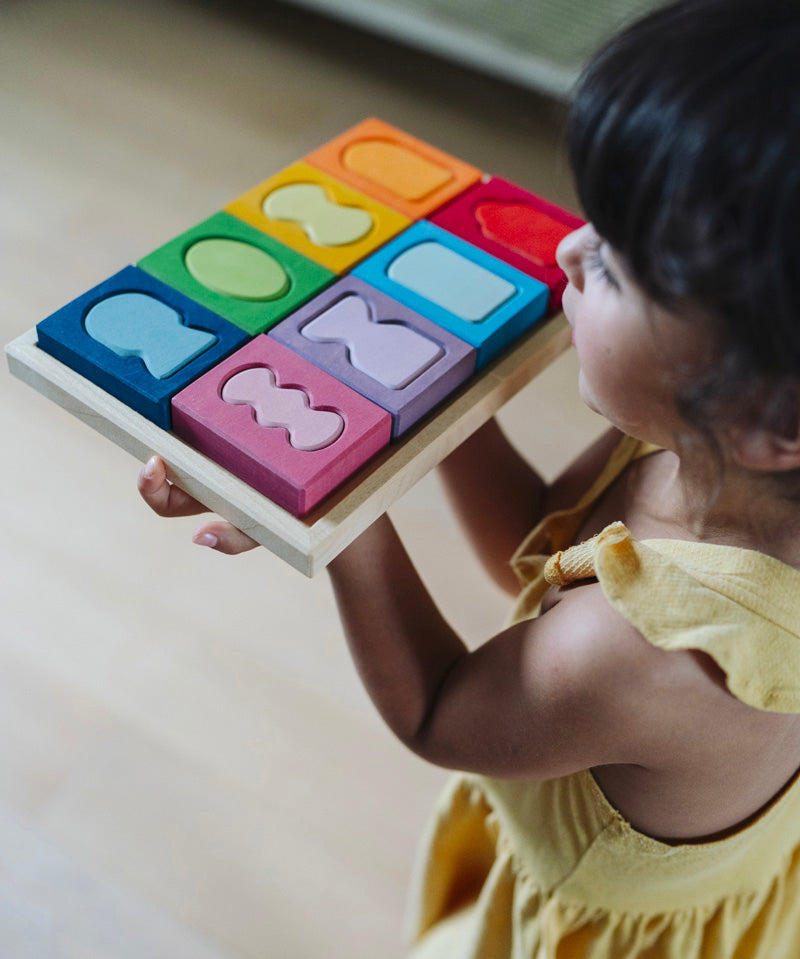 A child holding the Grimm's windows building set blocks set on the floor in front of them. These Grimm's toys are part of a wide range of wooden toys available here at Babipur. 
