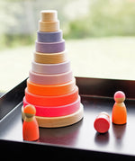 A set of 3 Grimms neon pink wooden friend dolls standing next to a large pink conical stacking tower on a black metal surface