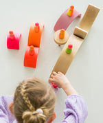 A top down view of a child playing with the Grimms wooden neon friend peg dolls on top of pink stacking rainbow pieces