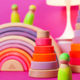 Grimms handcrafted wooden neon green peg doll standing in front of a neon pink stacking tower and rainbow