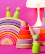 Grimms handcrafted wooden neon green peg doll standing in front of a neon pink stacking tower and rainbow