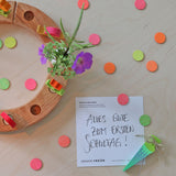 A flatlay of the green and pink neon birthday cone decorative figures next to the mixed neon confetti dots  and a celebration ring on a wooden surface 