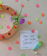 A flatlay of the green and pink neon birthday cone decorative figures next to the mixed neon confetti dots  and a celebration ring on a wooden surface 