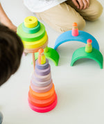 A close up of children playing with the Grimms wooden neon collection showing the pink and green conical tower, neon rainbow pieces with 3 neon friends on top