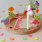 A Grimms wooden neon confetti dots scattered next to a celebration ring with different decorative figures on top including a purple singing bird, a orange flower, pink neon birthday cone, mushroom, snail, white vase and a beeswax candle. 