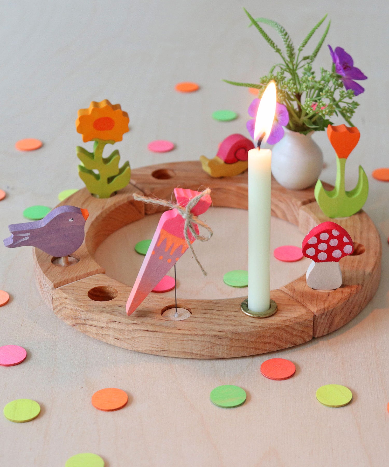 A Grimms wooden neon confetti dots scattered next to a celebration ring with different decorative figures on top including a purple singing bird, a orange flower, pink neon birthday cone, mushroom, snail, white vase and a beeswax candle. 