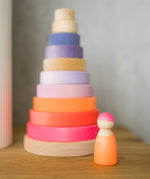 A Grimms neon pink wooden friend peg doll standing next to a conical stacking tower on a wooden surface
