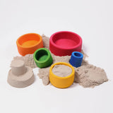 Grimms bright rainbow wooden sorting bowls with sand inside next to a small sandcastle