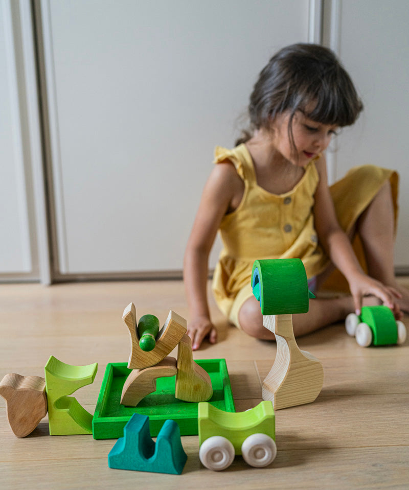 A child hand playing with the Grimm's green squirrel Building Set and green stacking cars. Wooden blocks in various shades of green and natural wood in a green coloured wooden storage tray. These Grimm's toys are part of a wide range of wooden toys available here at Babipur. 