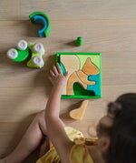 An overhead view of a child playing with the Grimm's green squirrel Building Set. Wooden blocks in various shades of green and natural wood in a green coloured wooden storage tray. These Grimm's toys are part of a wide range of wooden toys available here at Babipur. 