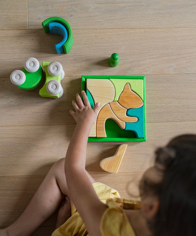 An overhead view of a child playing with the Grimm's green squirrel Building Set. Wooden blocks in various shades of green and natural wood in a green coloured wooden storage tray. These Grimm's toys are part of a wide range of wooden toys available here at Babipur. 