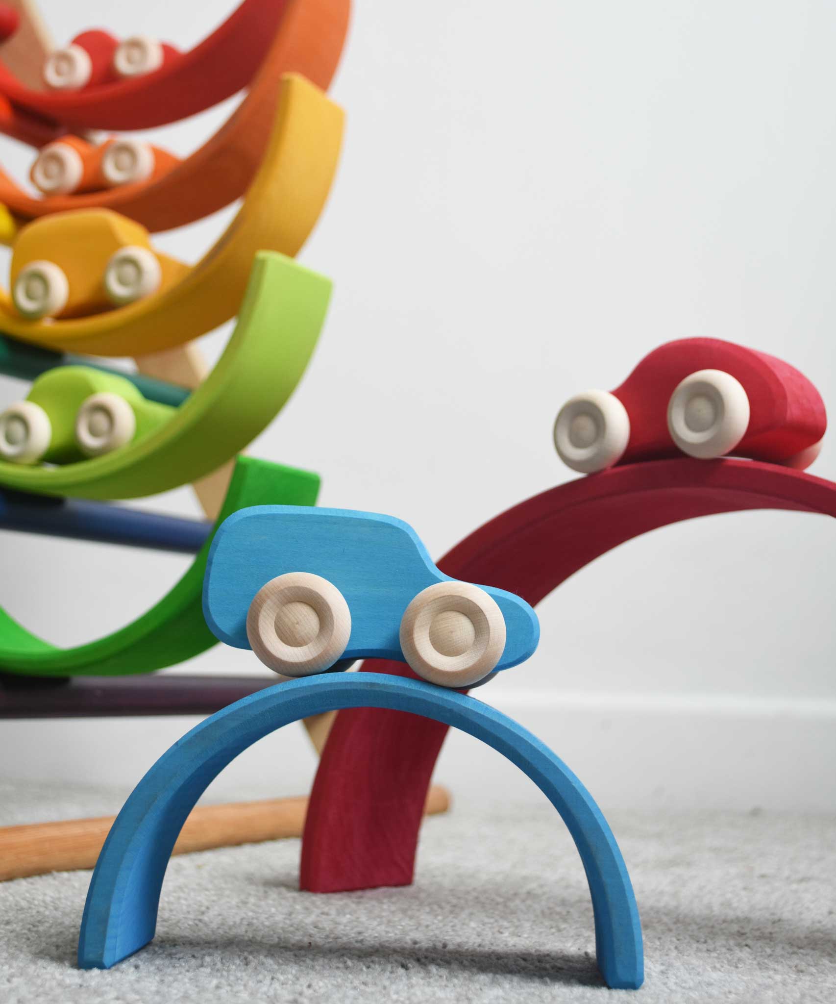 The cars from the Grimm's 6 Coloured Wooden Cars set placed on matching coloured pieces from the large Grimm's rainbow tunnel. The blue car can be seen at the front balanced on the blue coloured rainbow arch. These wooden toy cars are part of a wide range of Grimm's wooden toys available here at Babipur.
