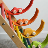 Four cars from the Grimm's 6 Coloured Wooden Cars set placed on matching coloured pieces from the large Grimm's rainbow tunnel. The rainbow arches have been balanced on the rungs of the Triclimb climbing frame. These wooden toy cars are part of a wide range of Grimm's wooden toys available here at Babipur.