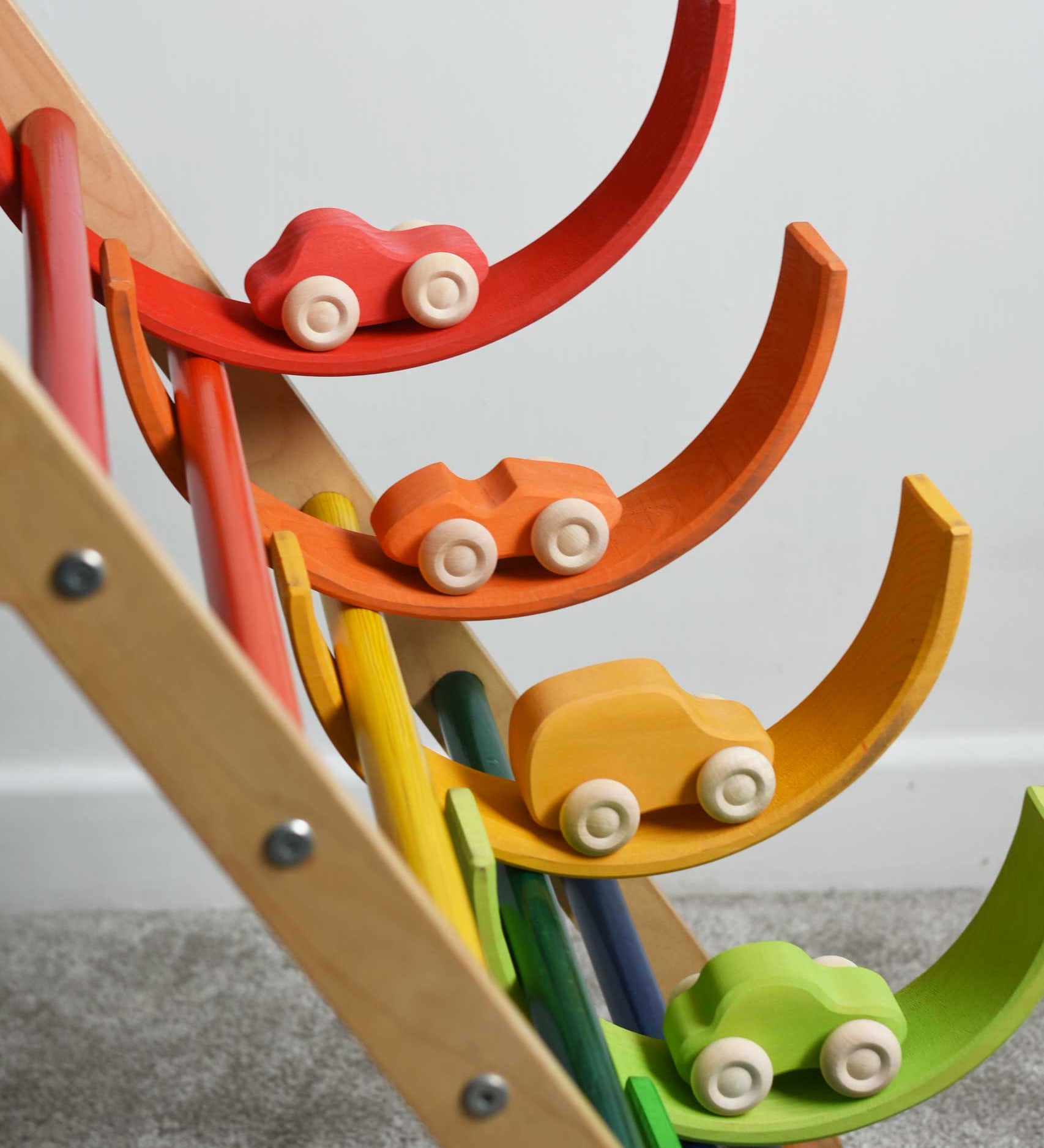 Four cars from the Grimm's 6 Coloured Wooden Cars set placed on matching coloured pieces from the large Grimm's rainbow tunnel. The rainbow arches have been balanced on the rungs of the Triclimb climbing frame. These wooden toy cars are part of a wide range of Grimm's wooden toys available here at Babipur.