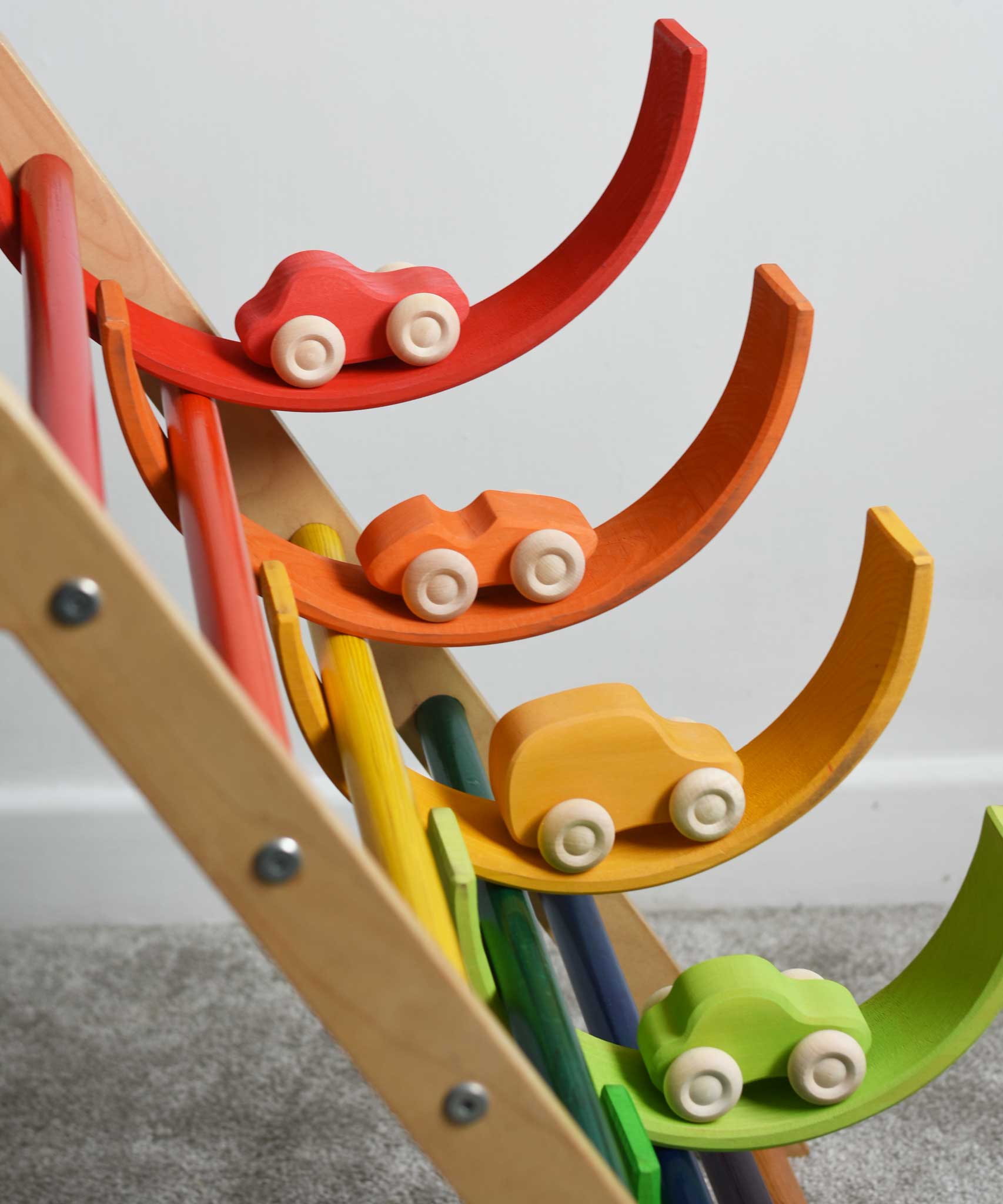 Four cars from the Grimm's 6 Coloured Wooden Cars set placed on matching coloured pieces from the large Grimm's rainbow tunnel. The rainbow arches have been balanced on the rungs of the Triclimb climbing frame. These wooden toy cars are part of a wide range of Grimm's wooden toys available here at Babipur.
