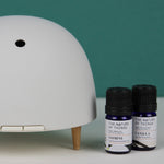 Maël Diffuser, a light grey dome plug-in essential oil diffuser with wooden feet, with two blue bottles of essential oils.