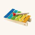 Haba Musical Metallophone, Xylophone Toy Instrument. A wooden drumming stick on top of a row of colourful flat metal bars pinned into a wooden base with each one being a different musical note, on a cream background