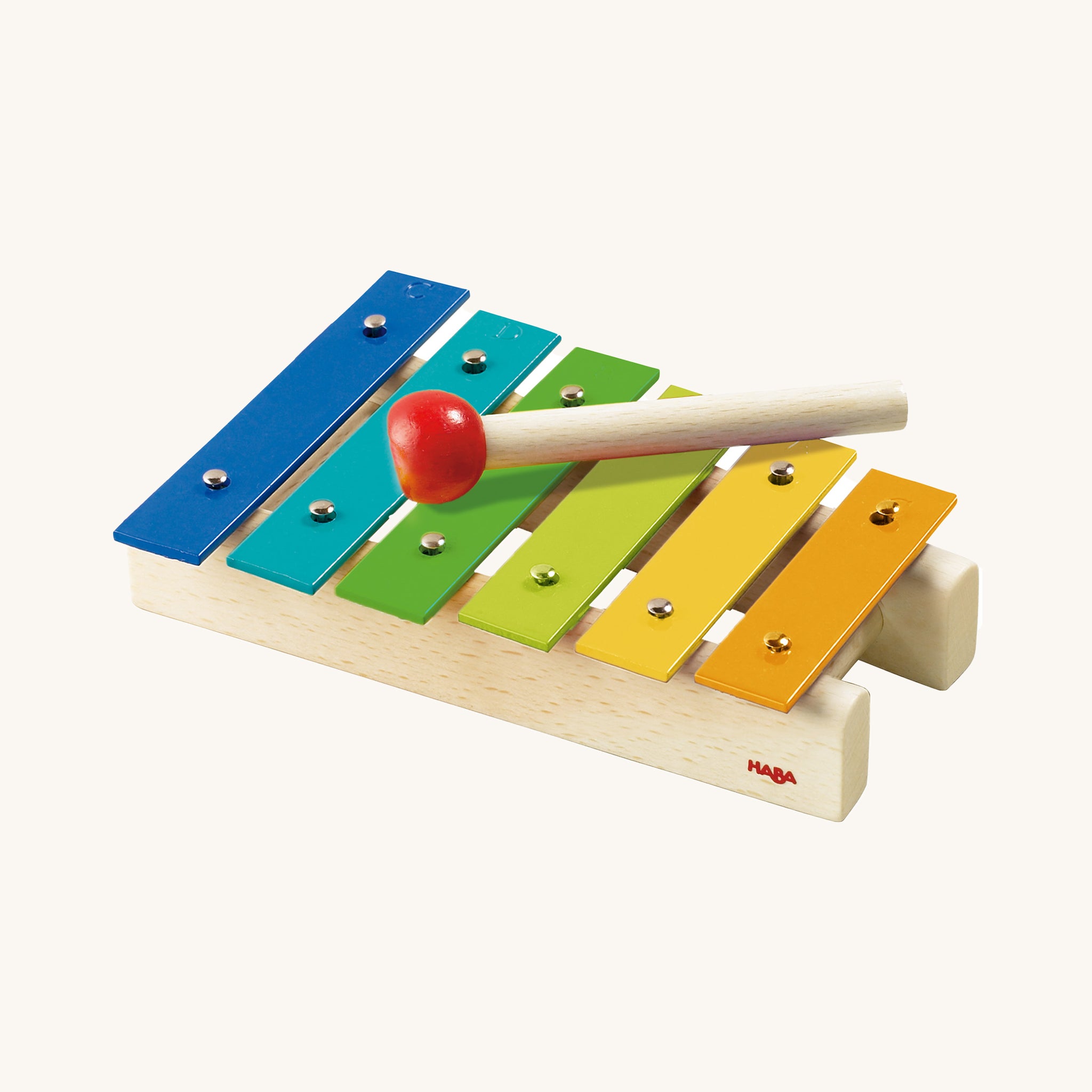 Haba Musical Metallophone, Xylophone Toy Instrument. A wooden drumming stick on top of a row of colourful flat metal bars pinned into a wooden base with each one being a different musical note, on a cream background
