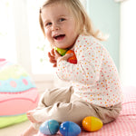 A child chewing on the blue Haba Musical Wooden Egg Toy whilst sat on a light blue rug with the other Haba Musical Eggs by their feet