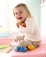 A child chewing on the blue Haba Musical Wooden Egg Toy whilst sat on a light blue rug with the other Haba Musical Eggs by their feet