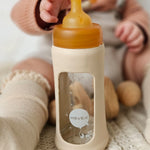 Baby sat on the floor reaching towards the Hevea Glass Bottle Wide Neck With Rubber Sleeve - 250ml in Sand