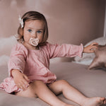 Young girl sat on a bed with a pink Hevea pacifier in her mouth