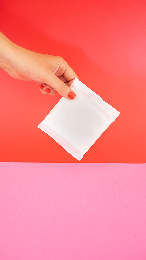 Hey Girls Natural Bamboo & Corn Fibre Disposable Day Pad wrapped in packaging held in someone's hand in front of a red and pink background