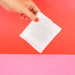 Hey Girls Natural Bamboo & Corn Fibre Disposable Day Pad wrapped in packaging held in someone's hand in front of a red and pink background