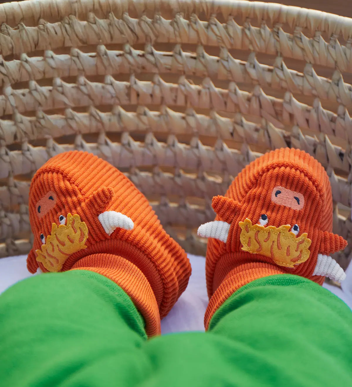 A close-up of a baby's feet wearing bright orange knit slippers shaped like cartoon monsters with yellow tufts, white tusks, and blue eyes, resting in a woven wicker basket.