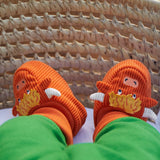 A close-up of a baby's feet wearing bright orange knit slippers shaped like cartoon monsters with yellow tufts, white tusks, and blue eyes, resting in a woven wicker basket.