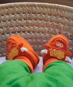 A close-up of a baby's feet wearing bright orange knit slippers shaped like cartoon monsters with yellow tufts, white tusks, and blue eyes, resting in a woven wicker basket.