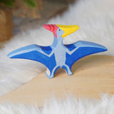 Holztiger Pteranodon pictured on a wooden play platform with white fluffy fabric behind. A blue flying dinosaur figure with handpainted details.