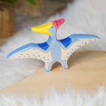 Holztiger Pteranodon pictured on a wooden play platform with white fluffy fabric behind. A blue flying dinosaur figure with handpainted details.