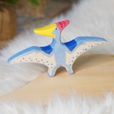 Holztiger Pteranodon pictured on a wooden play platform with white fluffy fabric behind. A blue flying dinosaur figure with handpainted details.