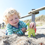 A child lying down on a sandy beach holding onto the Holztiger spinosaurus. The sky is blue and the sunshine is out