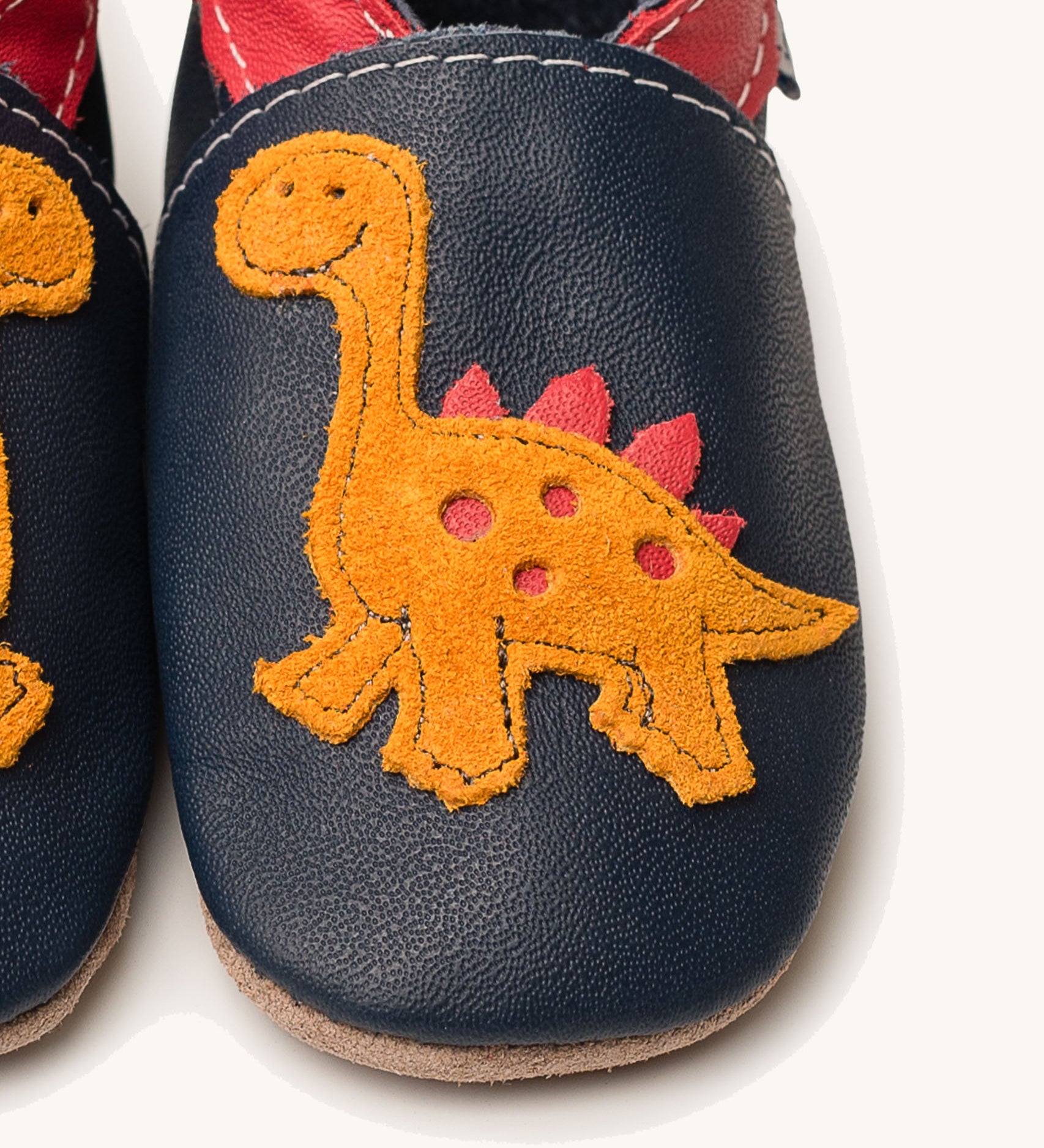 A close up of the Inch Blue navy dino baby shoes available at Babipur showing the orange suede dino cut out design
