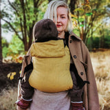 A woman babywearing a baby on her front using the Integra ochre yellow texture linen baby carrier available at a Babipur. Features a small hood for head protection.
