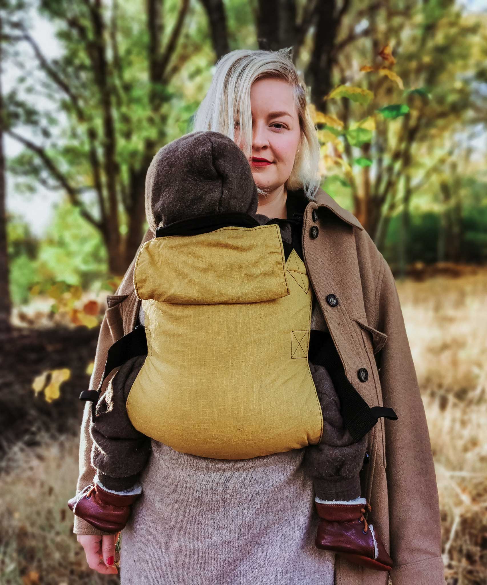 A woman babywearing a baby on her front using the Integra ochre yellow texture linen baby carrier available at a Babipur. Features a small hood for head protection.