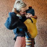 A woman babywearing a baby on her front using the Integra ochre yellow texture linen baby carrier available at a Babipur. Features a small hood for head protection.