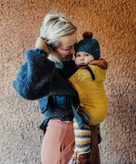 A woman babywearing a baby on her front using the Integra ochre yellow texture linen baby carrier available at a Babipur. Features a small hood for head protection.