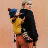 A woman babywearing a baby on her back using the Integra ochre yellow texture linen baby carrier available at a Babipur. Features a small hood for head protection.