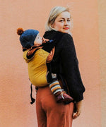 A woman babywearing a baby on her back using the Integra ochre yellow texture linen baby carrier available at a Babipur. Features a small hood for head protection.