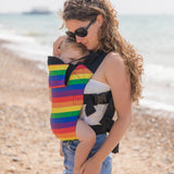 A mother walking on a beach carrying a baby in a rainbow design baby carrier from Integra available at Babipur.