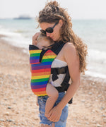 A mother walking on a beach carrying a baby in a rainbow design baby carrier from Integra available at Babipur.