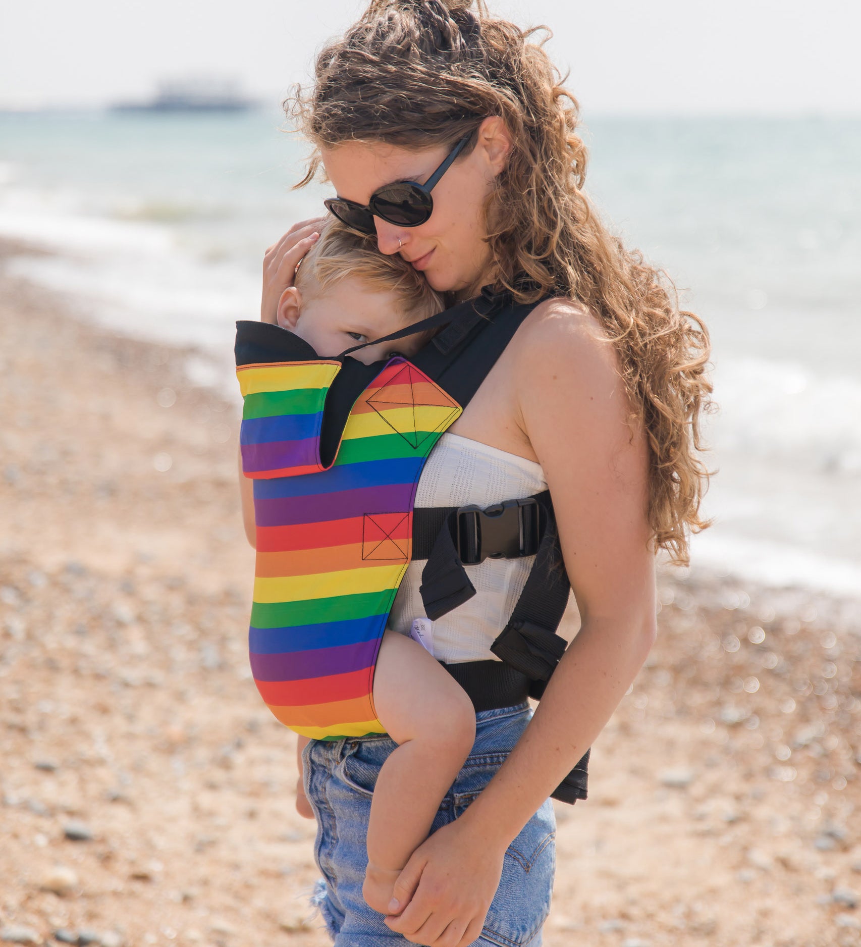 A mother walking on a beach carrying a baby in a rainbow design baby carrier from Integra available at Babipur.