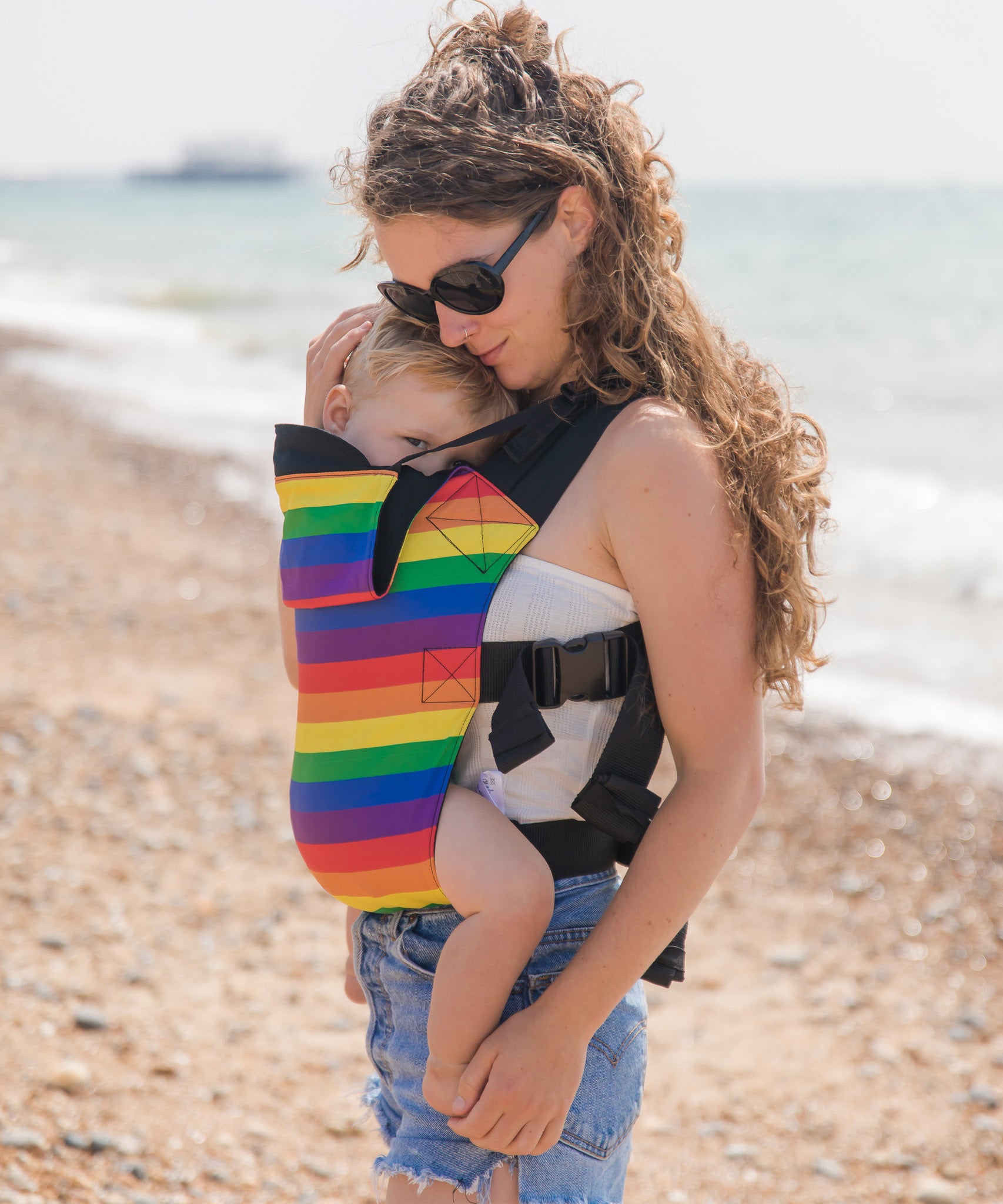A mother walking on a beach carrying a baby in a rainbow design baby carrier from Integra available at Babipur.