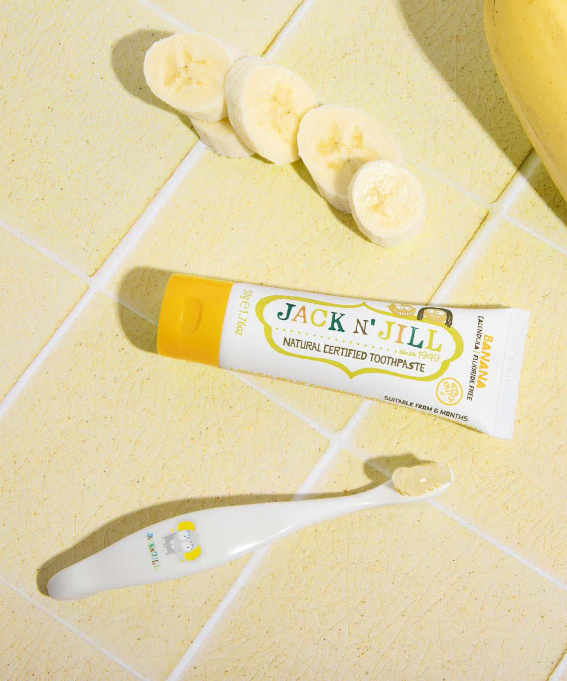 A 50g tube of Jack N' Jill banana flavoured fluoride free kid's toothpaste placed on a yellow tiled surface next to a toothbrush. 